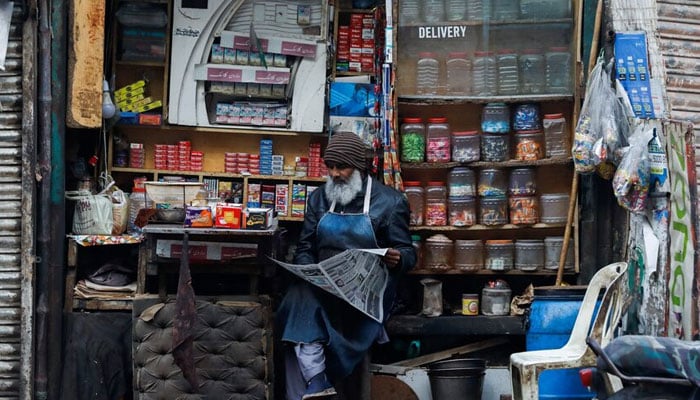 A man reads newspaper while selling betel leaves, known as pan, cigarettes and candies from a shop in Karachi. — Reuters/File