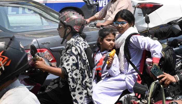 A woman rides a scooty with her children, while holding a mobile phone on a busy road after school in Lahore, on May 7, 2026. — APP