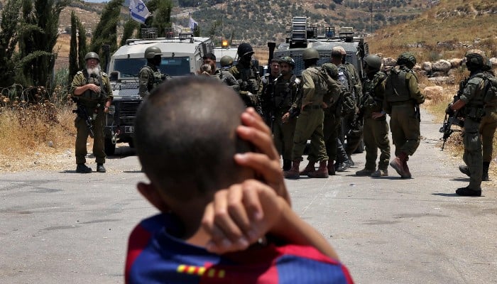 A Palestinian boy looks on as Israeli security forces stand guard before Palestinians gathering to protest threats of confiscation of their land in the Palestinian village of Idna, west of Hebron in the occupied West Bank, on June 12, 2026. — AFP
