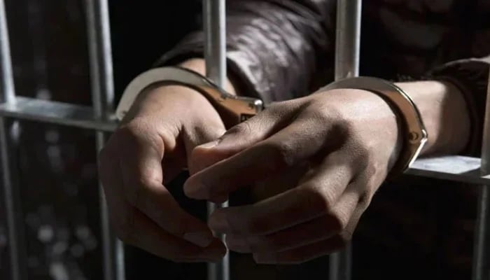 A representational image of a handcuffed man standing behind bars. — APP/File